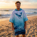 Poncho serviette à capuche motif dauphin turquoise, porté par un homme souriant, debout sur la plage au lever du soleil.