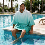 Poncho serviette à capuche dégradé menthe, porté par un homme assis au bord d'une piscine.