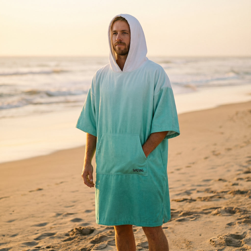Poncho serviette à capuche dégradé menthe, porté par un homme debout sur le sable au coucher du soleil.