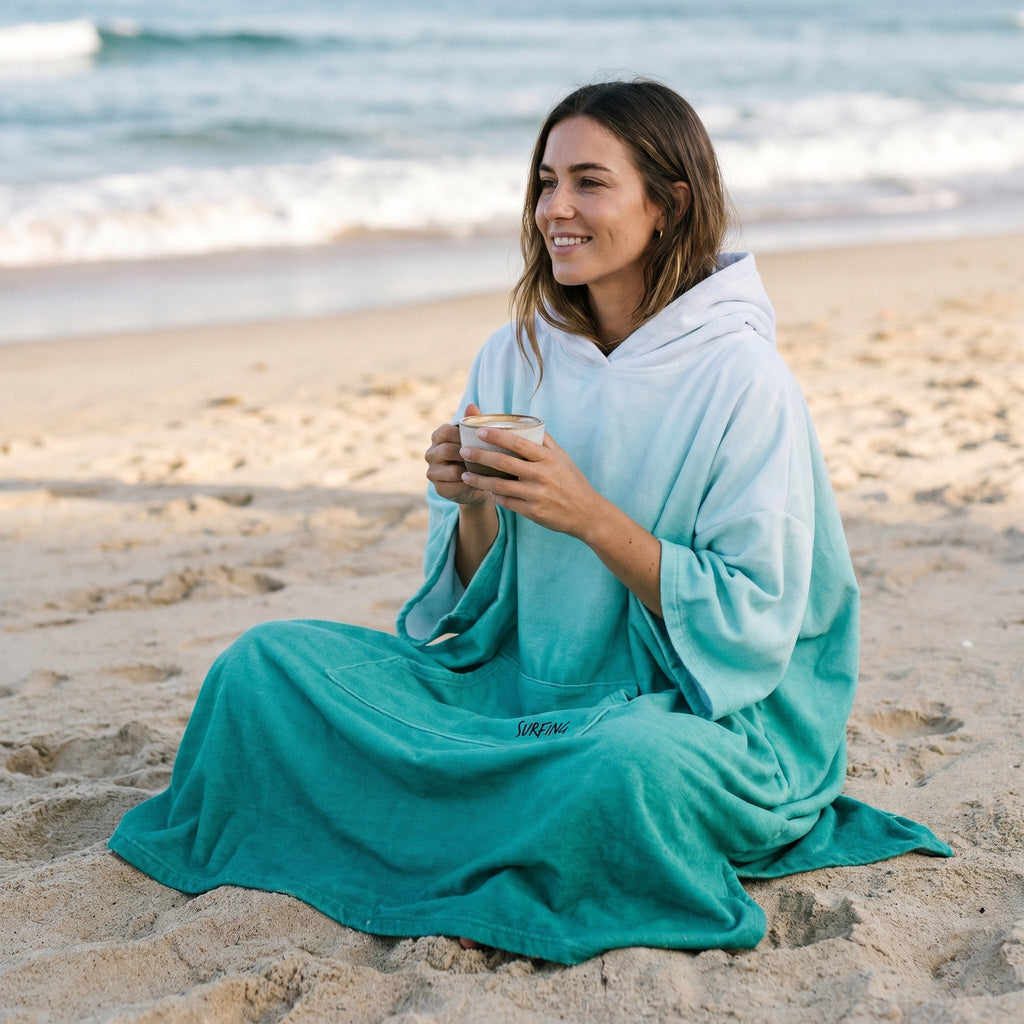 Poncho serviette à capuche dégradé menthe, porté par une femme assise sur la plage tenant une tasse.