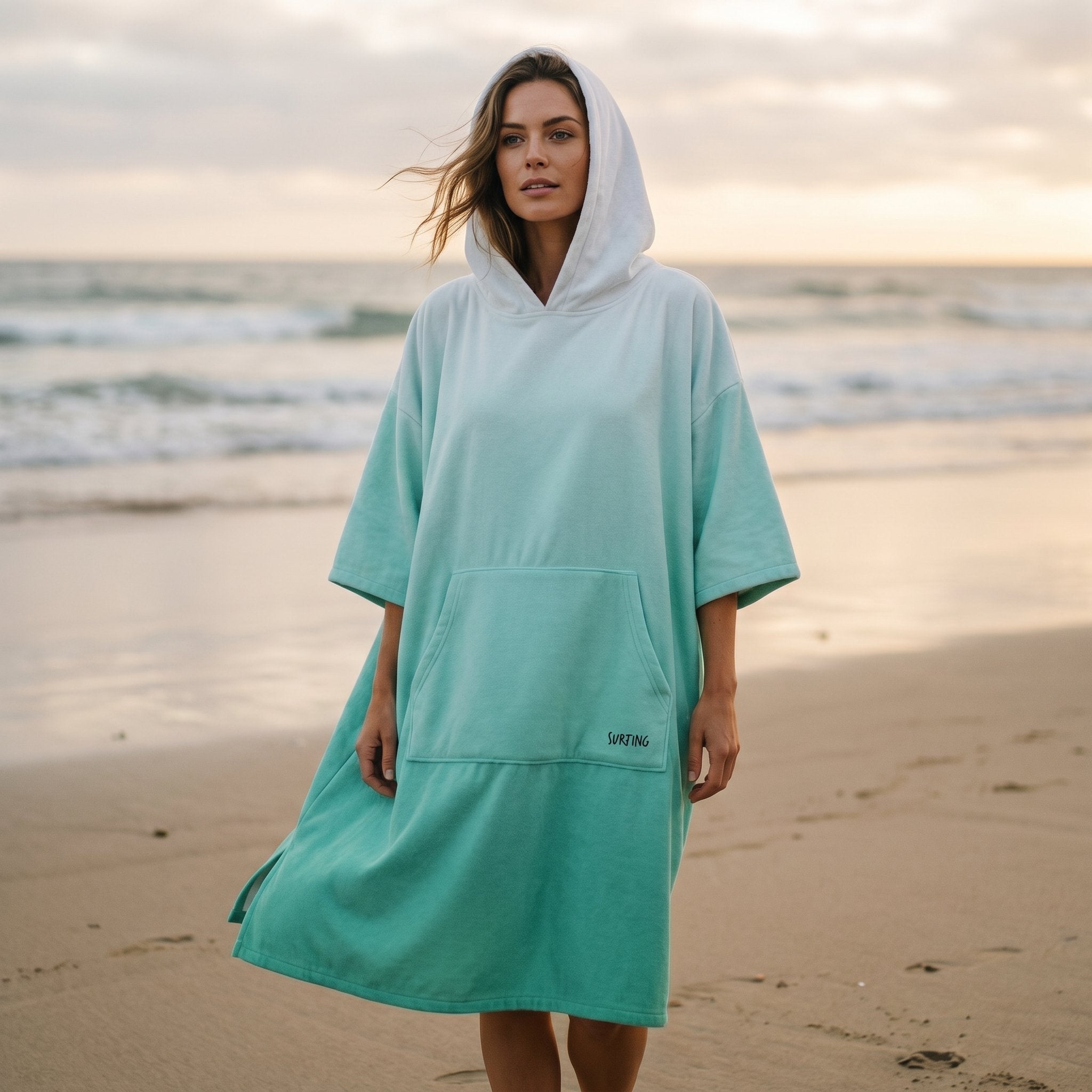 Poncho serviette à capuche dégradé menthe, porté par une femme debout sur le sable face à l'objectif.