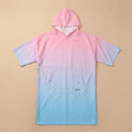 Poncho serviette à capuche dégradé rose et bleu, posé à plat sur un fond beige clair.