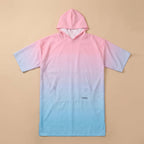 Poncho serviette à capuche dégradé rose et bleu, posé à plat sur un fond beige clair.