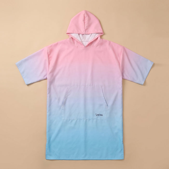 Poncho serviette à capuche dégradé rose et bleu, posé à plat sur un fond beige clair.