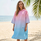 Poncho serviette à capuche dégradé rose et bleu, porté par une femme debout sur une plage de sable fin.