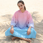 Poncho serviette à capuche dégradé rose et bleu, porté par une femme assise en tailleur sur le sable, vue de face.