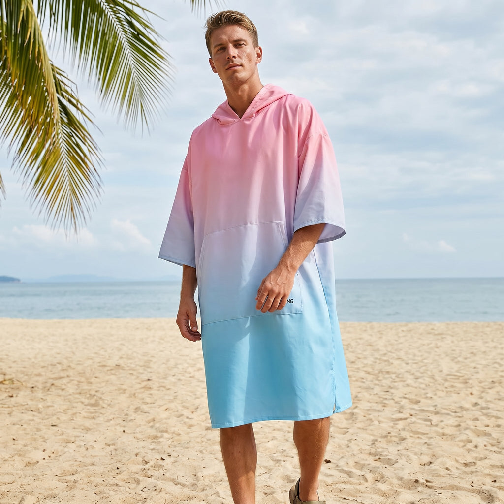 Poncho serviette à capuche dégradé rose et bleu, porté par un homme debout sur la plage, regardant vers l'avant.