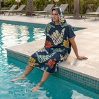 Poncho serviette à capuche motif shakas colorés, porté par un homme assis au bord d'une piscine, les pieds dans l'eau.