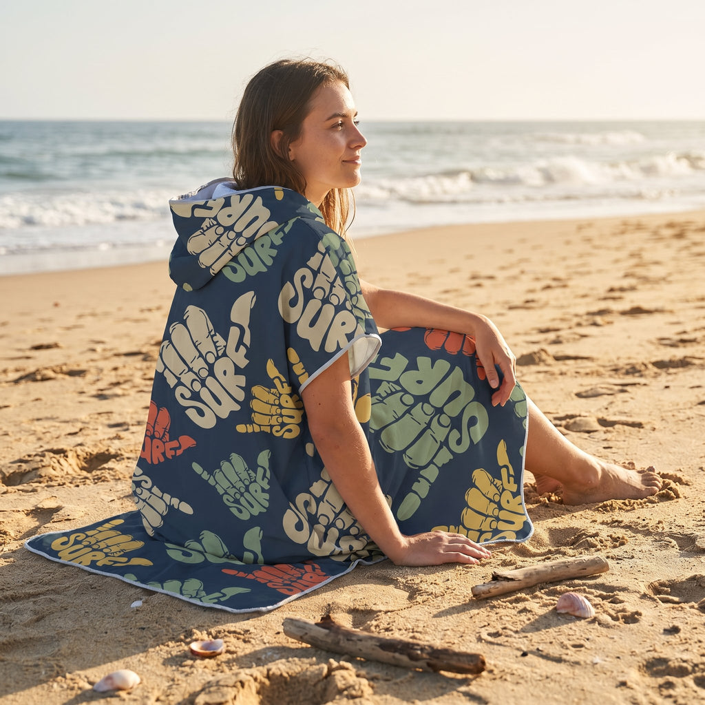 Poncho serviette à capuche motif shakas colorés, porté par une femme assise sur le sable, vue de côté.