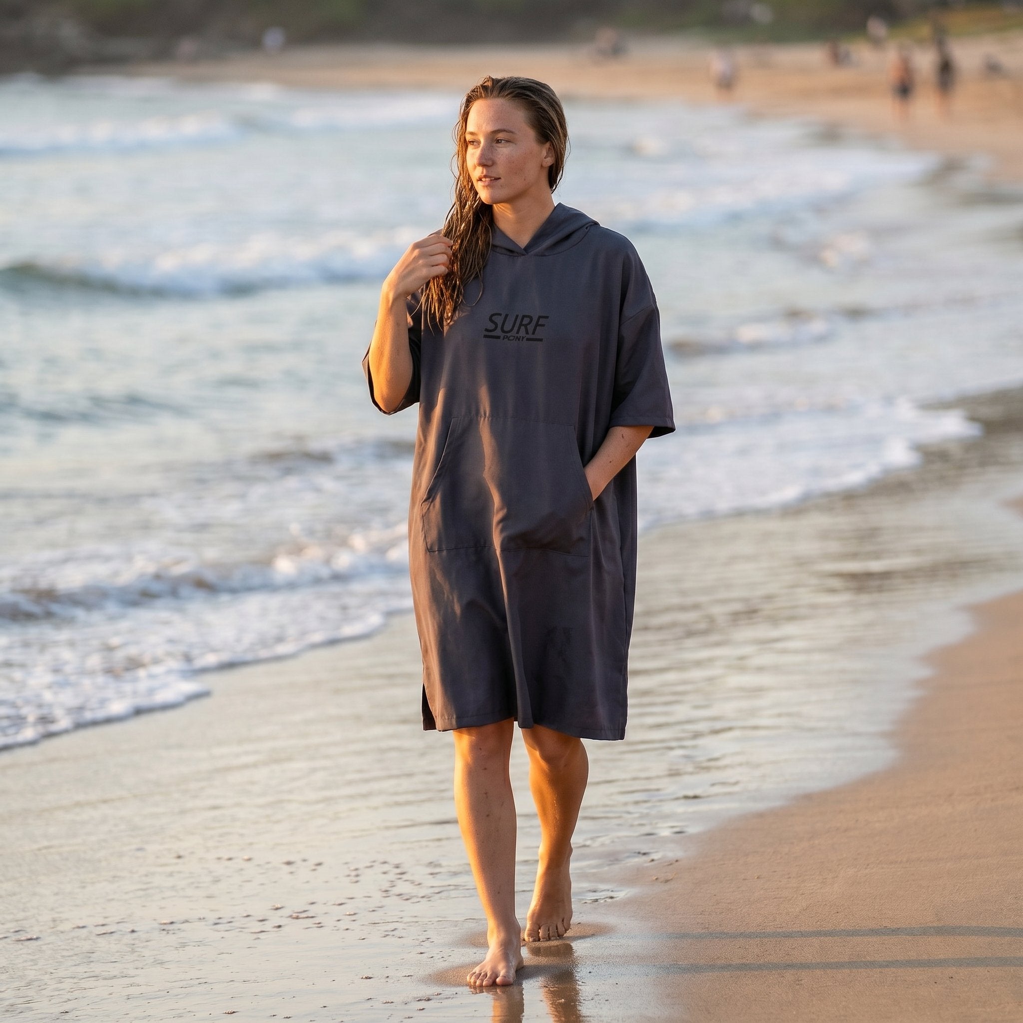 Poncho de surf à capuche taupe uni, porté par une femme marchant sur la plage au bord de l'eau.