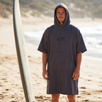 Poncho de surf à capuche taupe uni, porté par un homme debout sur le sable, près d'une planche de surf.