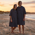 Poncho de surf à capuche taupe uni, porté par un homme et une femme sur la plage au coucher du soleil.