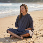 Poncho de surf à capuche taupe uni, porté par une femme assise en tailleur sur le sable de la plage.