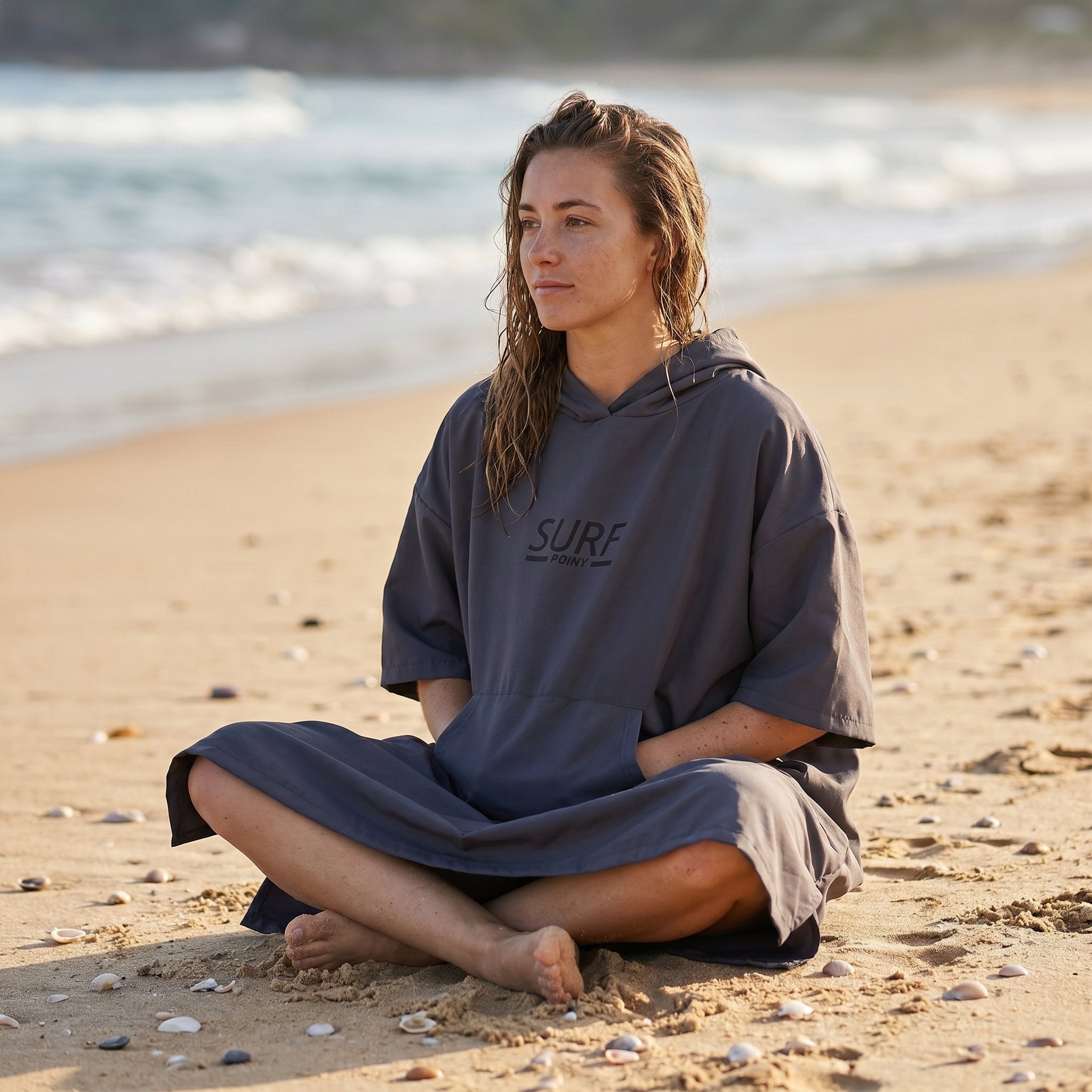 Poncho de surf à capuche taupe uni, porté par une femme assise en tailleur sur le sable de la plage.