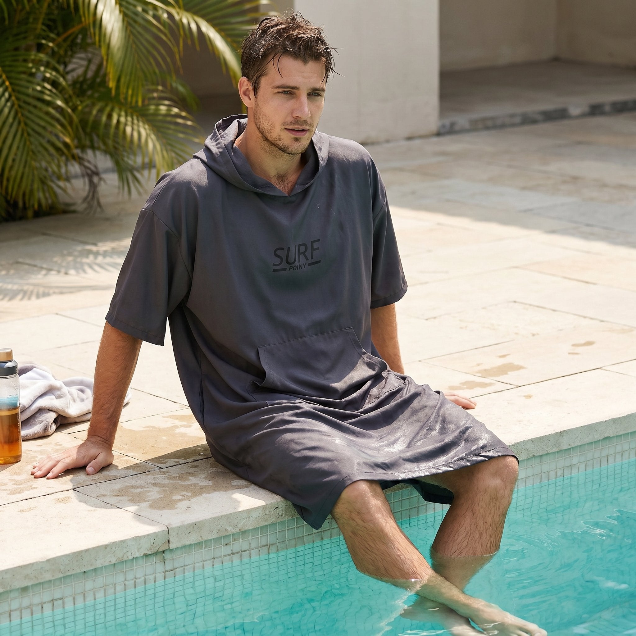 Poncho de surf à capuche taupe uni, porté par un homme assis au bord d'une piscine, les pieds dans l'eau.