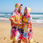 Poncho serviette a capuche motif tropical rose et jaune, porté par un couple souriant sur la plage.