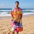 Poncho serviette a capuche motif tropical rose et jaune, porté par un homme debout sur la plage.