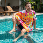 Poncho serviette a capuche motif tropical rose et jaune, porté par un homme assis au bord d'une piscine.