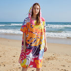 Poncho serviette a capuche motif tropical rose et jaune, porté par une femme debout face à l'océan.