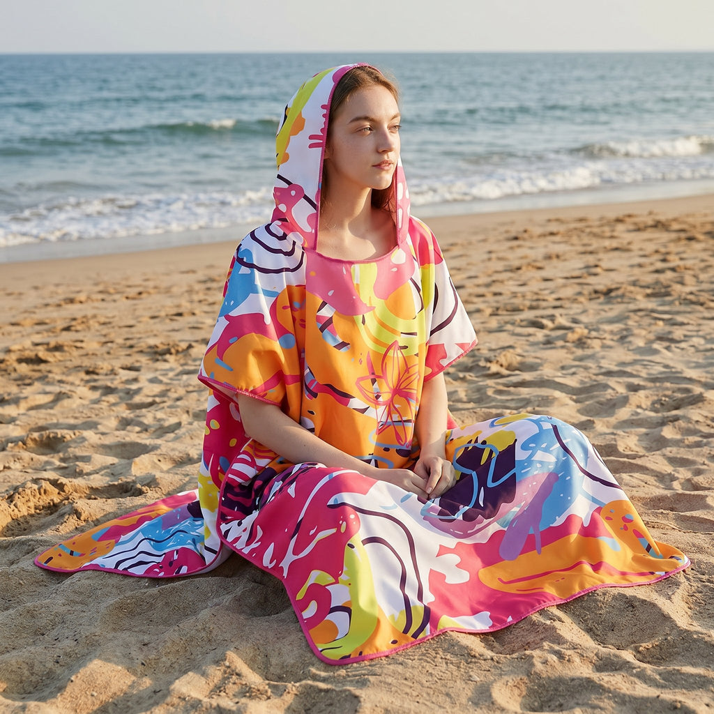Poncho serviette a capuche motif tropical rose et jaune, porté par une femme assise sur le sable.