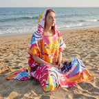 Poncho serviette a capuche motif tropical rose et jaune, porté par une femme assise sur le sable.