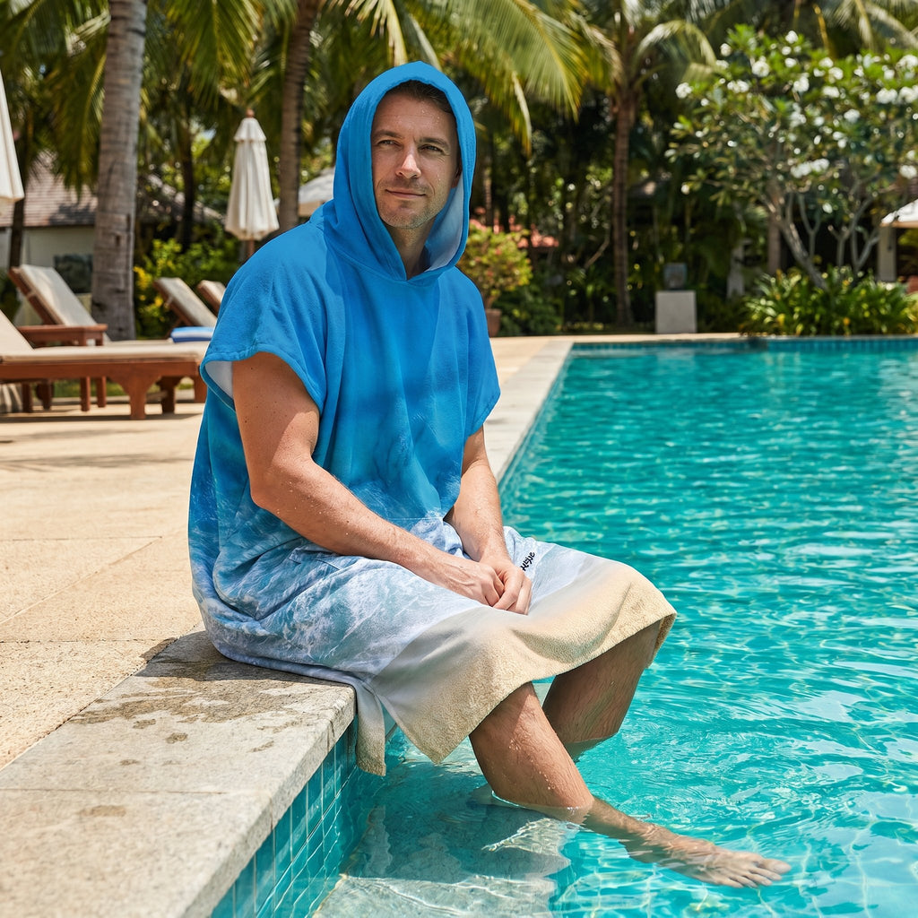 Poncho serviette à capuche motif vague océan, porté par un homme assis au bord d'une piscine.