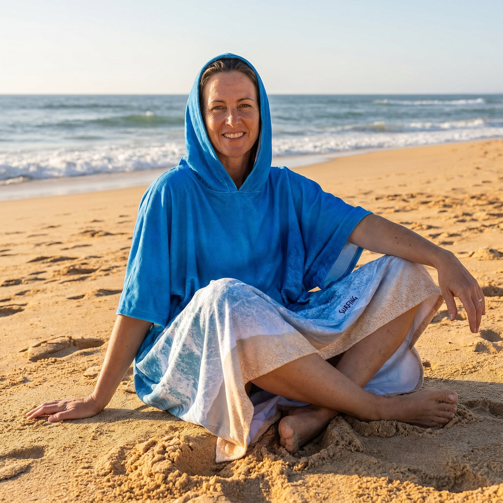 Poncho serviette à capuche motif vague océan, porté par une femme assise en tailleur sur le sable.
