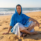 Poncho serviette à capuche motif vague océan, porté par une femme assise en tailleur sur le sable.