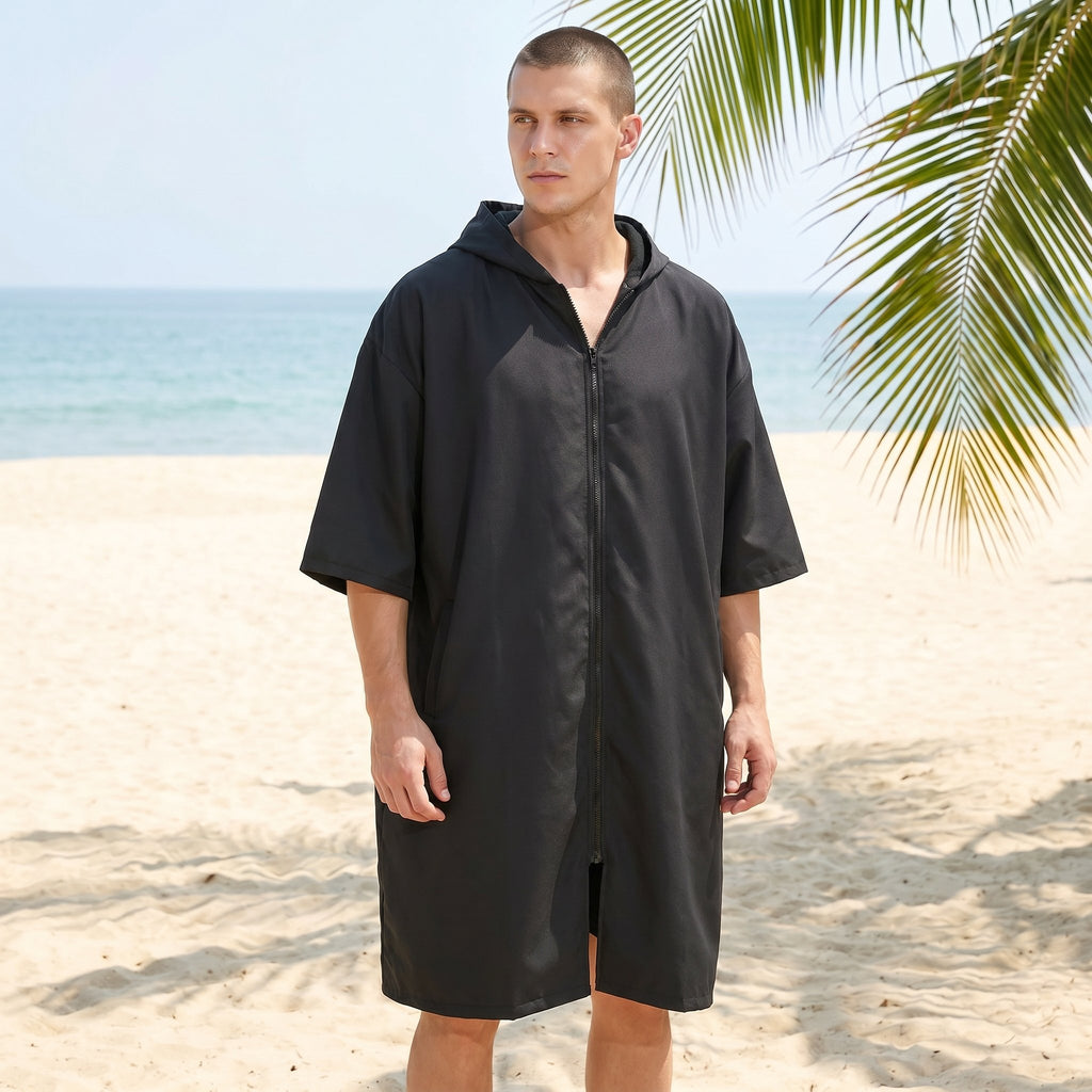 Poncho serviette à capuche noir uni, porté par un homme sur une plage ensoleillée, face à l'océan.