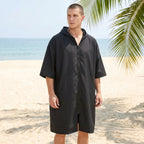 Poncho serviette à capuche noir uni, porté par un homme sur une plage ensoleillée, face à l'océan.