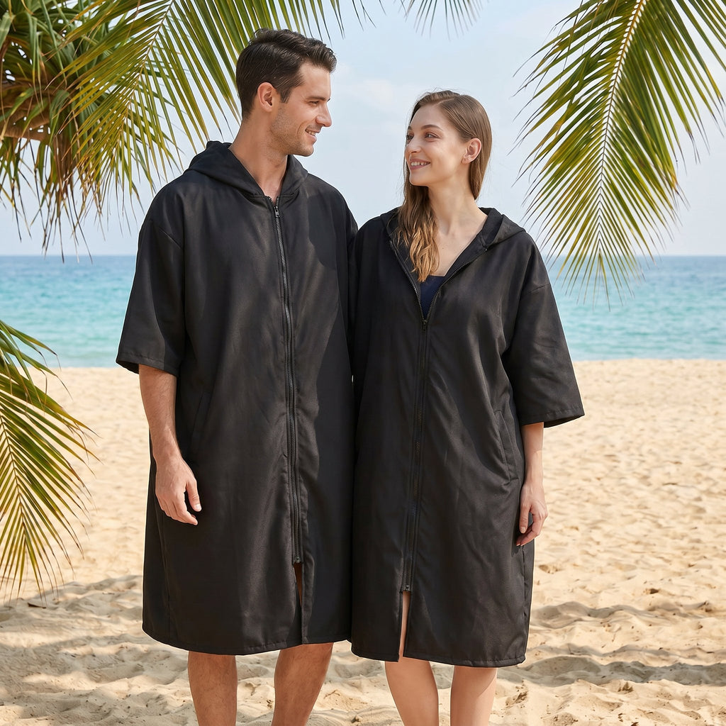 Poncho serviette à capuche noir uni, porté par un couple souriant sur le sable, avec la mer en arrière-plan.