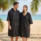 Poncho serviette à capuche noir uni, porté par un couple souriant sur le sable, avec la mer en arrière-plan.