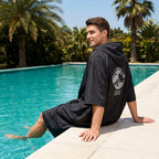 Poncho serviette à capuche noir uni, porté par un homme assis au bord d'une piscine, les pieds dans l'eau.