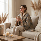 Poncho plaid · Carreaux beiges, porté par une femme souriante assise sur un canapé, avec une tasse fumante.