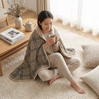 Poncho plaid · Carreaux beiges, porté par une femme assise en tailleur sur un tapis, buvant une tasse.