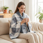 Poncho polaire à carreaux gris et blanc porté par une femme souriante assise sur un canapé, tenant une tasse.
