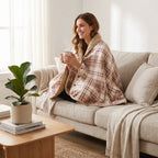 Poncho polaire à carreaux rosés porté par une femme souriante, assise sur un canapé, tenant une tasse fumante.