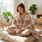 Poncho polaire à carreaux rosés porté par une femme assise en tailleur sur un coussin de sol, buvant une boisson chaude.