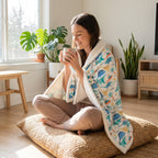Poncho polaire à capuche motif dinosaures, porté par une femme assise en tailleur sur des coussins.