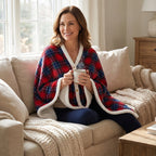 Poncho polaire tartan rouge et bleu porté par une femme assise sur un canapé, tenant une tasse fumante, vue de face.