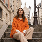 Poncho pull femme col roulé orange, porté par une femme assise sur des marches en pierre.