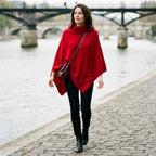 Poncho pull femme col roulé rouge, porté par une femme marchant au bord de l'eau avec un pont en arrière-plan.