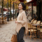 Poncho pull femme col roulé sable, porté par une femme souriante de profil devant une terrasse de café.