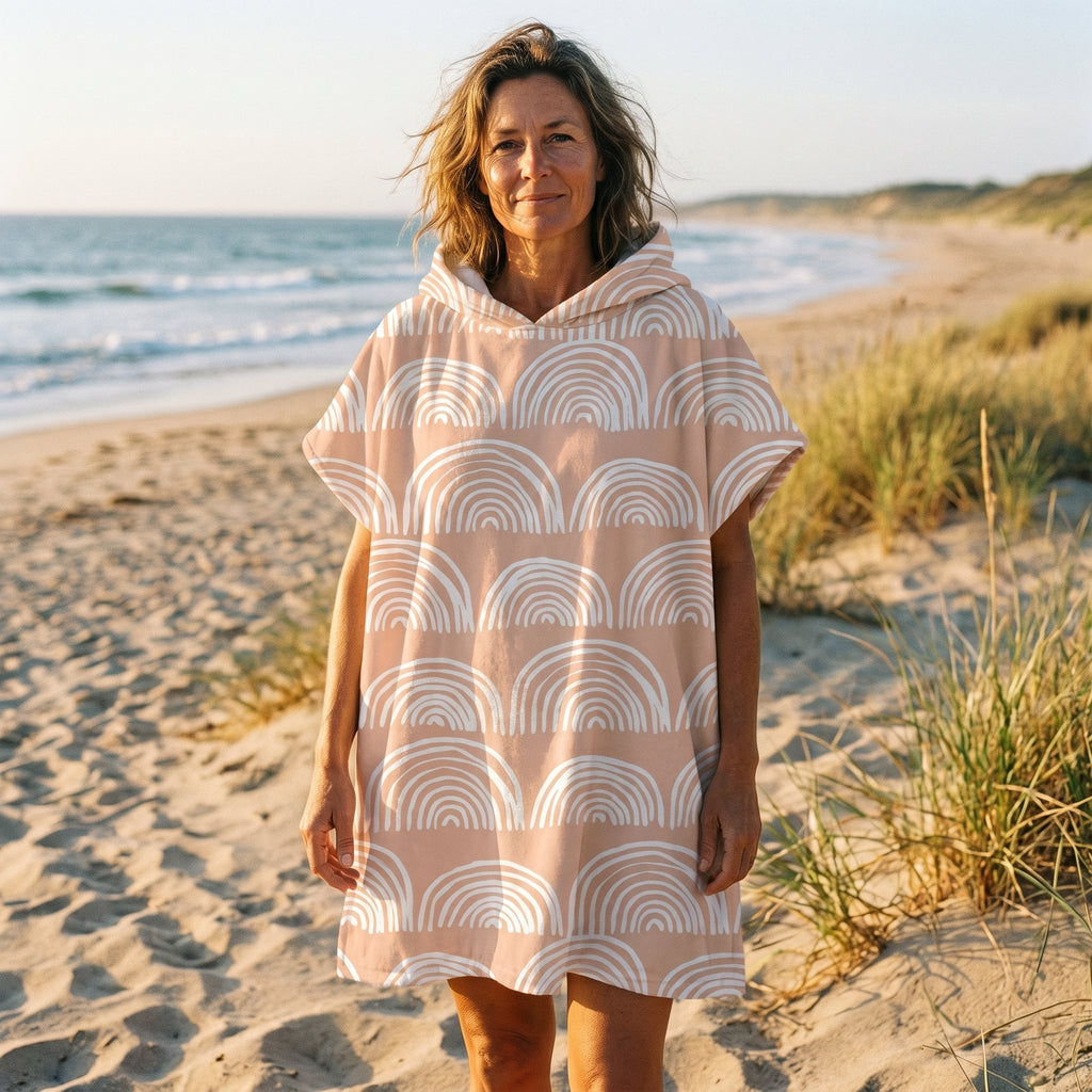 Poncho serviette a capuche motif Arches pêche, porté par une femme souriante, debout sur le sable face à l'océan.