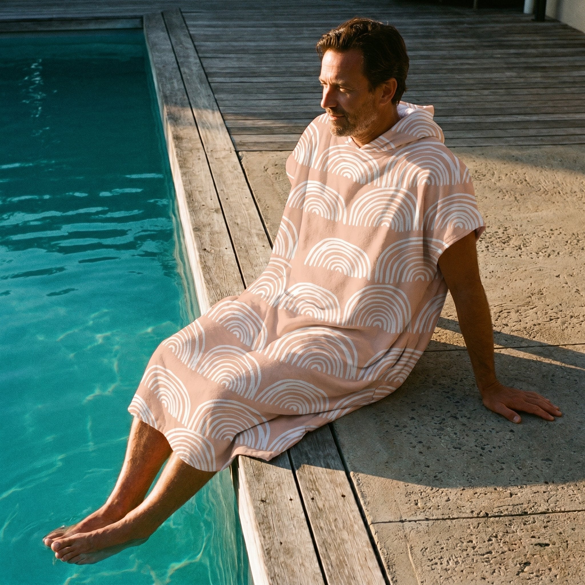 Poncho serviette a capuche motif Arches pêche, porté par un homme assis au bord d'une piscine, les pieds dans l'eau.
