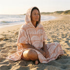Poncho serviette a capuche motif Arches pêche, porté par une femme assise en tailleur sur le sable, face à la mer.