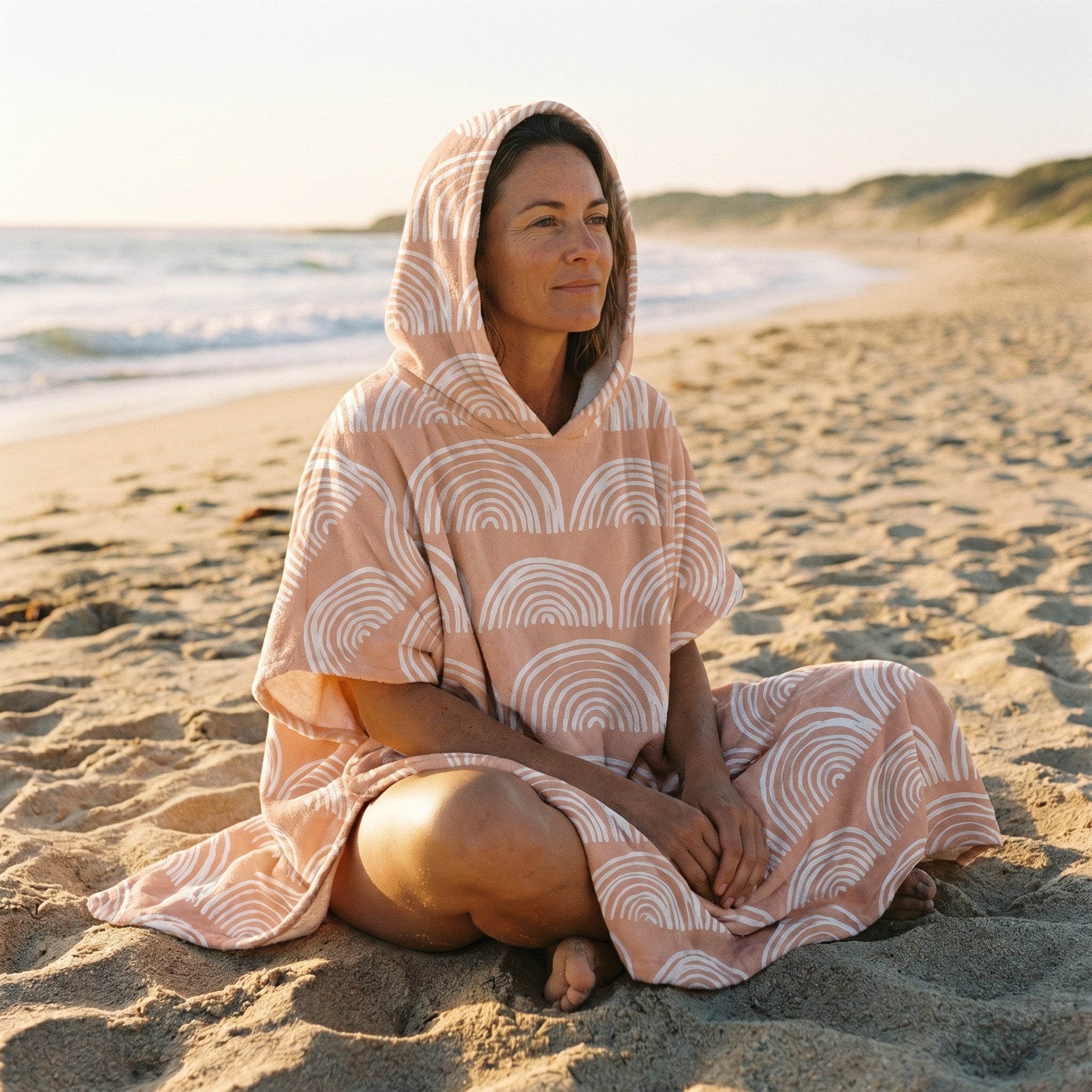 Poncho serviette a capuche motif Arches pêche, porté par une femme assise en tailleur sur le sable, face à la mer.