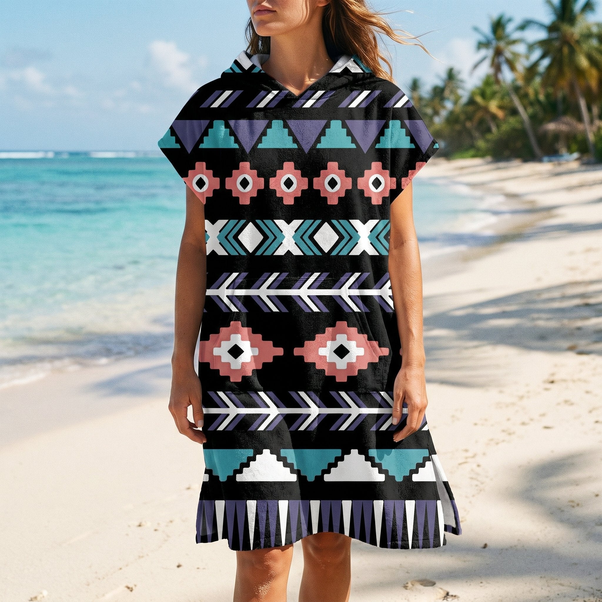 Poncho serviette a capuche motif aztèque rose et turquoise, porté par une femme, vue de face sur une plage ensoleillée.