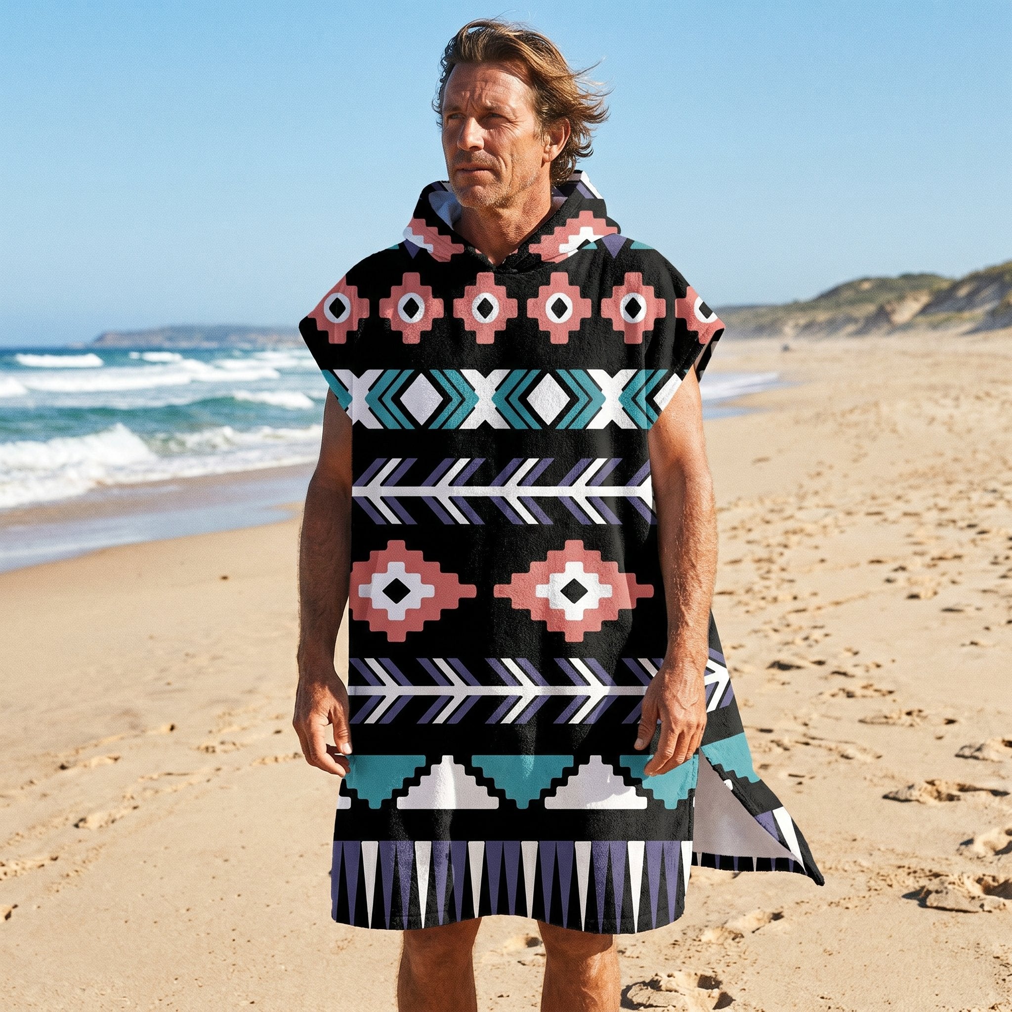 Poncho serviette a capuche motif aztèque rose et turquoise, porté par un homme, en plan large sur le sable d'une plage.