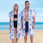 Poncho serviette à capuche motif coquillages bleus, porté ouvert par un couple souriant sur une plage ensoleillée.